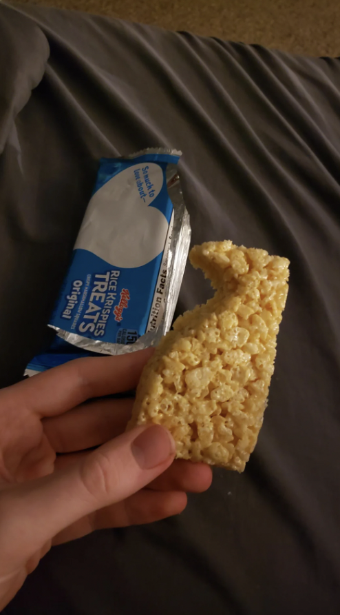 A hand holding a partially unwrapped Rice Krispies Treats bar over a dark gray bedspread. The packaging is blue and white, with the visible text indicating "Rice Krispies Treats" and "Original." The treat appears partially eaten.