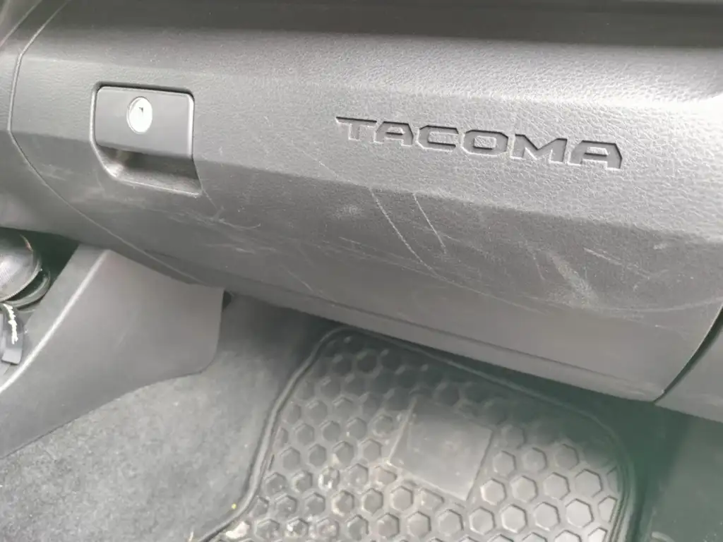 Close-up of a black car's glove compartment with the word "TACOMA" embossed on it. The area around the gloves compartment shows some scratches. Below, a black rubber floor mat with a hexagonal pattern is visible.