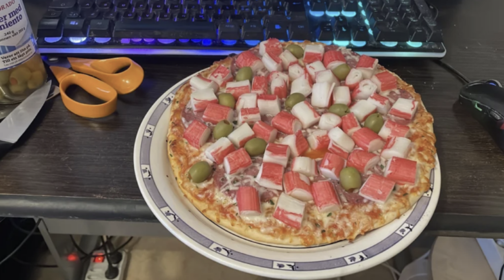 An image of a frozen pizza that has imitation crab and whole green olives on it. 