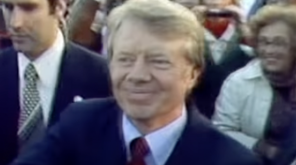 A man in a suit and red tie is smiling and shaking hands with people in a crowd, reminiscent of the warmth found in Jimmy Carter quotes. Another man in a suit stands nearby, while a woman in a light jacket is visible in the background. The scene appears to be outdoors.