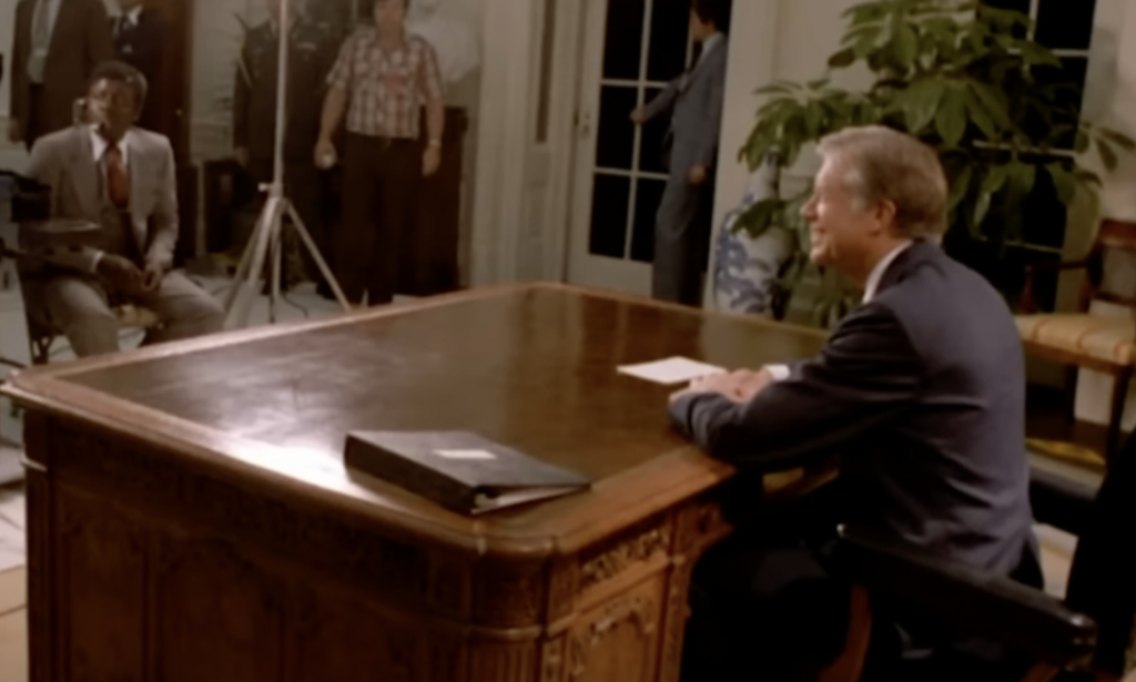 A man in a suit sits at an ornate wooden desk in an office, preparing to speak or be interviewed. A few people, including a cameraman with equipment, bustle in the background setting up. The room features a large plant and a door leading outside. Nearby, someone is quoting Jimmy Carter as part of the setup.