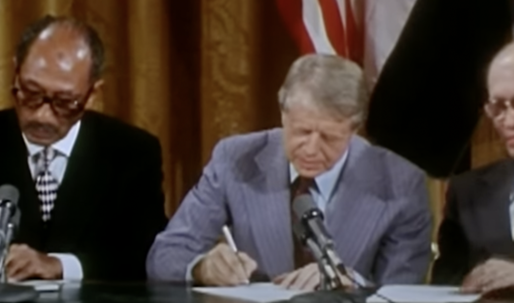 Three men are seated at a table, with the man in the center signing a document. Dressed in formal attire, they have microphones placed in front of them. An American flag is partially visible in the background, evoking a sense of patriotism reminiscent of famous Jimmy Carter quotes on unity and leadership.