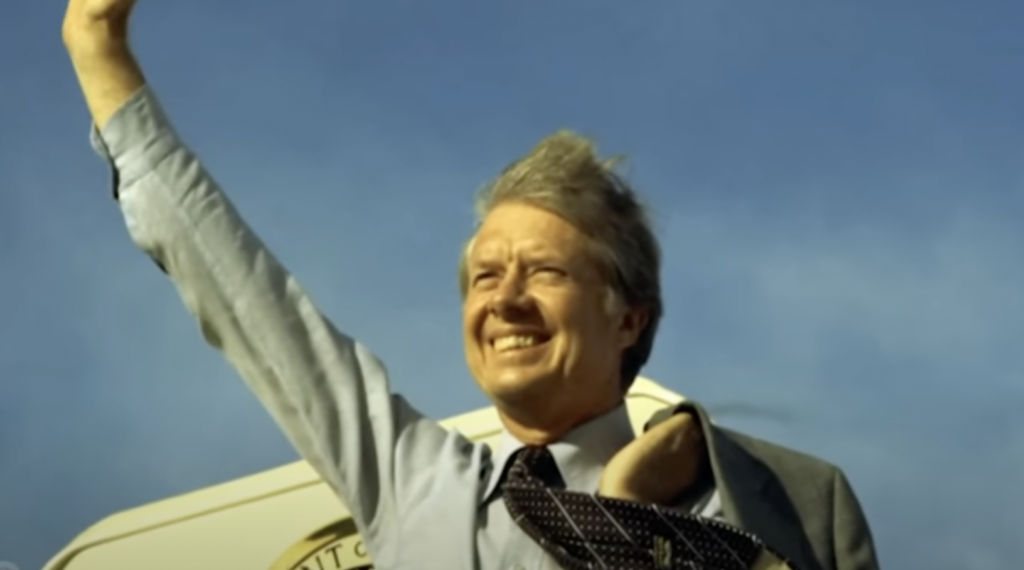 A man in a suit and tie stands outside with a backdrop of blue sky and part of an airplane. He is smiling and raising his right arm in a wave, while his left hand appears to hold items against his chest. His hair is being blown by the wind, invoking the spirit of one of those timeless Jimmy Carter quotes.