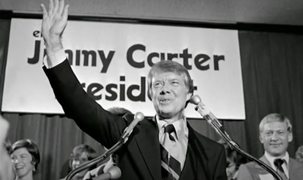 Black-and-white photo of a man in a suit waving with his right hand and smiling while standing behind multiple microphones. He is on stage with other people in the background, and a banner that reads "Jimmy Carter President" is visible behind him, evoking famous Jimmy Carter quotes about leadership.