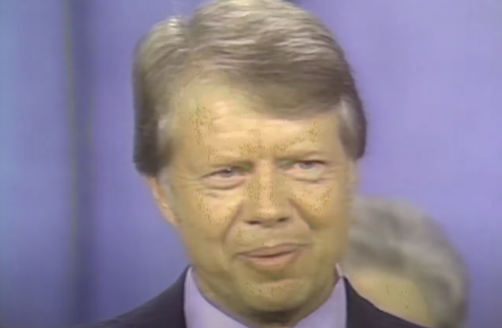 A person with light brown hair and a light complexion stands in front of a blue background, wearing a dark-colored suit and light-colored shirt. Smiling gently, the serene expression brings to mind one of those timeless Jimmy Carter quotes about kindness and perseverance.