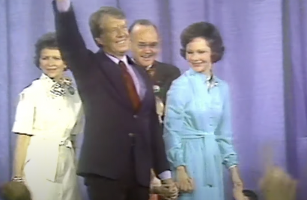 A man in a suit waves happily while standing alongside three people in front of a purple curtain. Two women in dresses and another man in a suit smile and look on. The group appears joyous, possibly inspired by one of Jimmy Carter's quotes, and is likely on a stage at a public event.