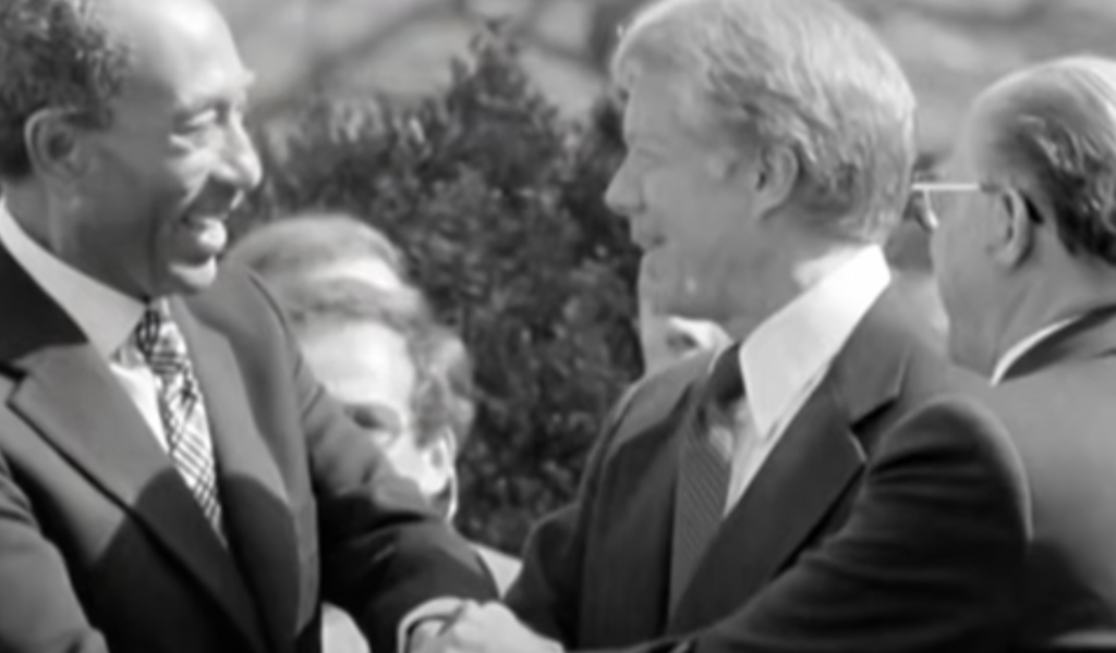 A black-and-white photo of three men, dressed in suits and ties, engaged in a friendly conversation. The two men on the left are shaking hands and smiling warmly at each other, perhaps exchanging Jimmy Carter quotes. The third man, on the right, is partially visible and also smiling.