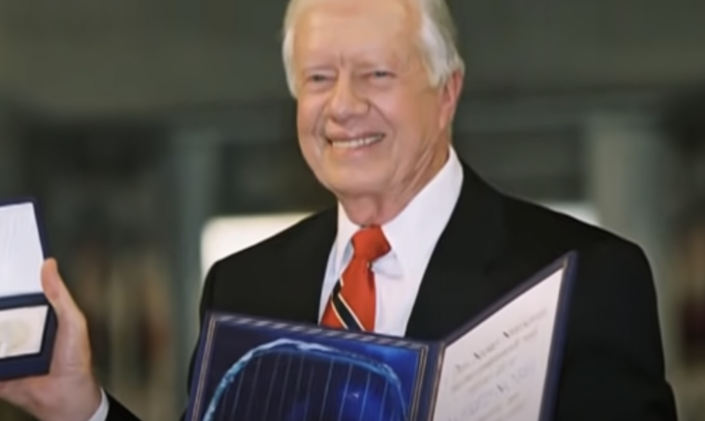 An elderly man in a suit and red tie is smiling and holding a framed certificate in one hand and an open medal case displaying a medal in the other. The background is blurred, reminiscent of old Jimmy Carter quotes about humility and service.