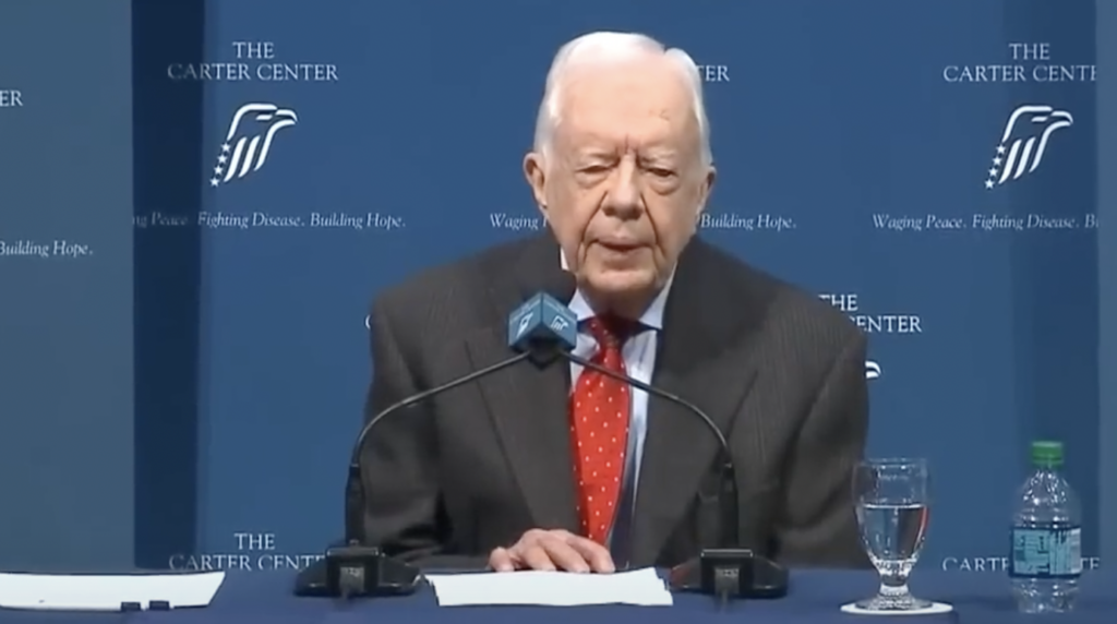 An elderly man with white hair and wearing a dark suit with a red tie speaks into a microphone. Seated at a table with a water bottle and glass of water in front of him, he is framed by a blue banner featuring "THE CARTER CENTER" text and logos. The scene evokes an air of reflection, reminiscent of famous Jimmy Carter quotes.