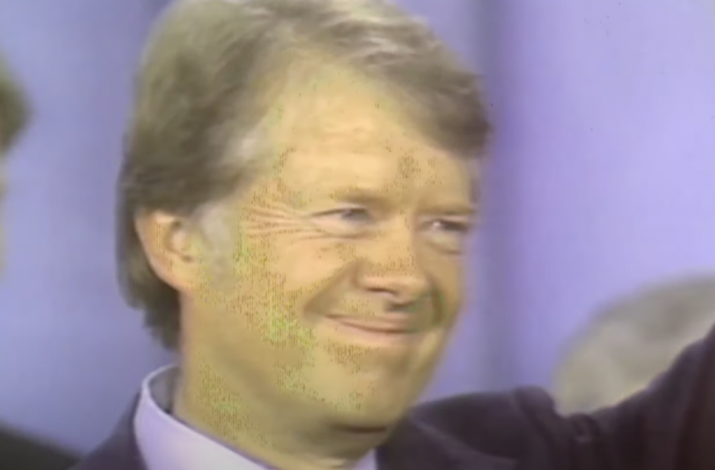 A man with light hair and a suit smiles and waves with one arm raised, reminiscent of jimmy carter quotes. The background is out of focus with light blue and gray tones.