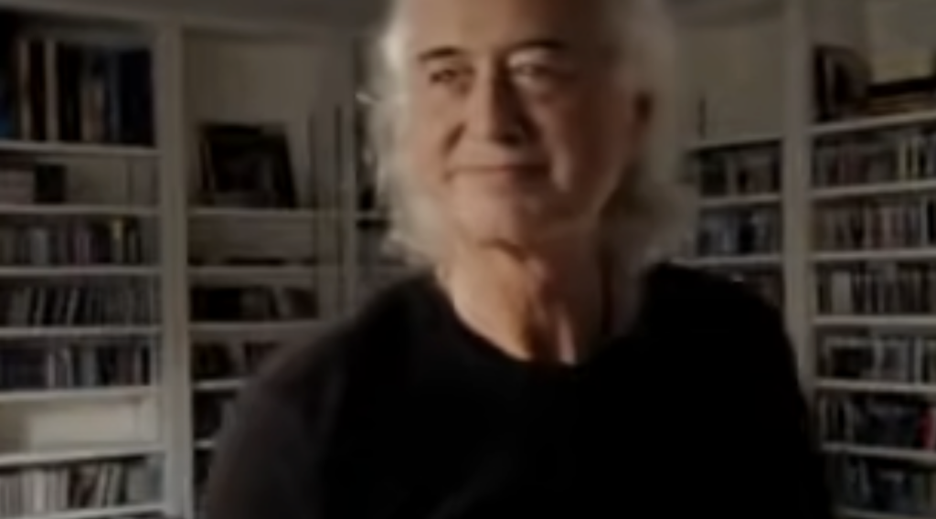 An older man with long gray hair and a black shirt stands in front of shelves filled with books and CDs. He is looking off to the side, smiling slightly. The background appears to be a cozy room, softly lit.