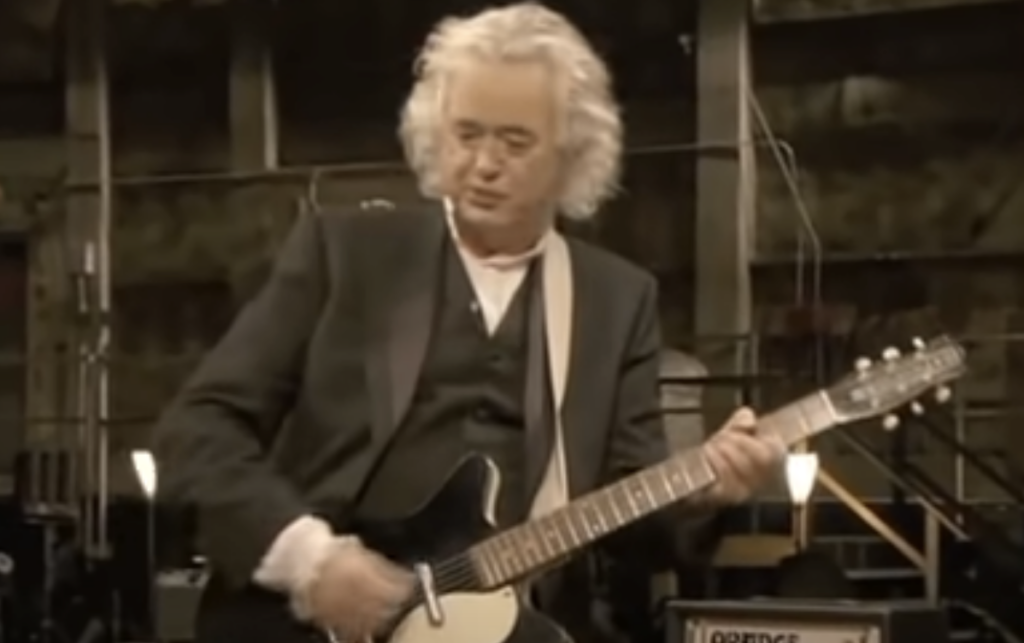 A person with shoulder-length gray hair, wearing a black suit and white shirt, is playing an electric guitar. The background shows a dimly lit industrial-like setting with metal beams and equipment.