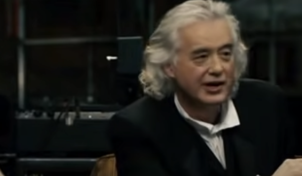 A person with long, gray hair and dressed in a dark collared shirt and jacket is speaking. They appear to be in a dimly lit indoor setting, possibly a studio or interview setting, with various equipment or instruments in the background.