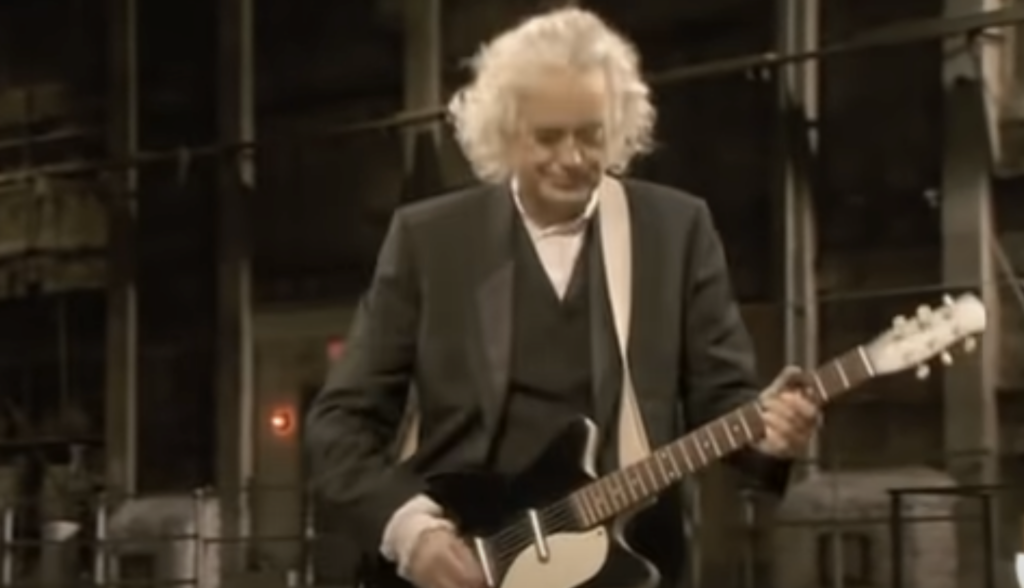 An older person with white hair is playing an electric guitar. They are wearing a black jacket over a white shirt. The background appears to be an industrial or warehouse setting with muted lighting.