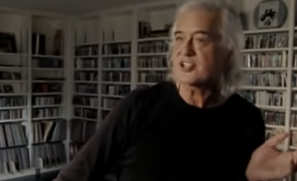 A person with long white hair is speaking and gesturing with one hand. They are wearing a black shirt and are standing in a room filled with bookshelves that appear to hold CDs or DVDs. The room has a bright, well-lit atmosphere.
