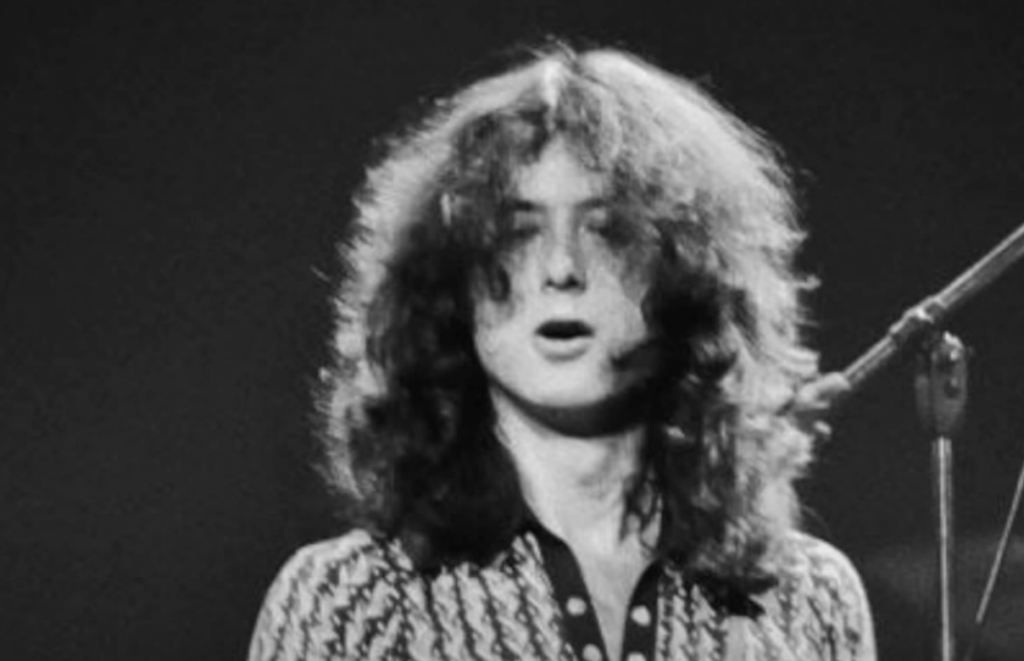 A black and white photograph of a person with long, wavy hair performing on stage, singing into a microphone. The person is wearing a patterned shirt and appears to be engaged in their performance.