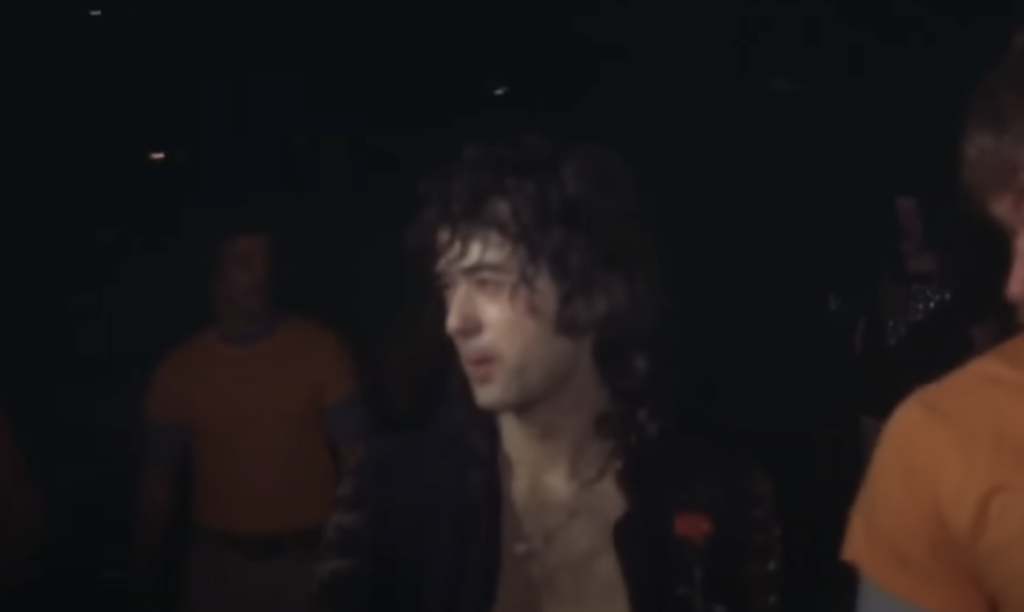 A person with long, dark, curly hair is seen in a dimly lit environment. The individual is wearing a black outfit, has a focused expression, and slightly wet face likely due to perspiration. Other people in orange shirts are visible in the background.