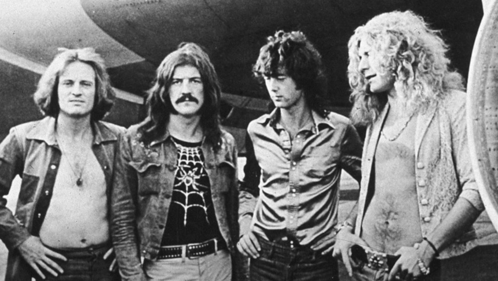 Four men with long hair stand in a line, posing outdoors. They appear to be part of a rock band, dressed in casual 1970s fashion, with some wearing open shirts and jackets. They are standing in front of what looks like an airplane or large vehicle.