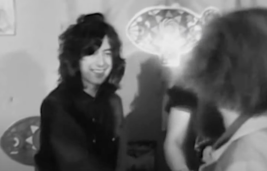 A black-and-white image of three people in a room. Two people with long hair are shaking hands and smiling. Another person is in the background. The scene is lit by a light fixture on the wall behind them. A small section of a poster is visible on the left.