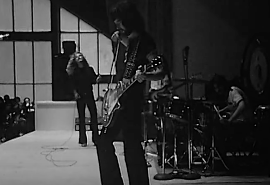 A black and white photo of a rock band performing on stage. The guitarist is in the foreground, playing and looking down. Behind him, the lead vocalist is singing near a microphone stand, and the drummer is partially visible on the right side of the image.