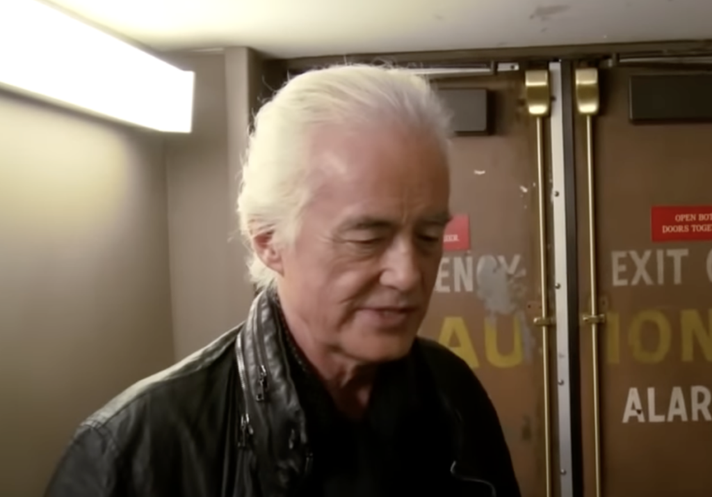 An elderly man with long, white hair is dressed in a black leather jacket and dark shirt. He is standing indoors in front of a pair of wooden doors with warning signs on them. The lighting in the room is bright, casting a soft glow on his face and hair.