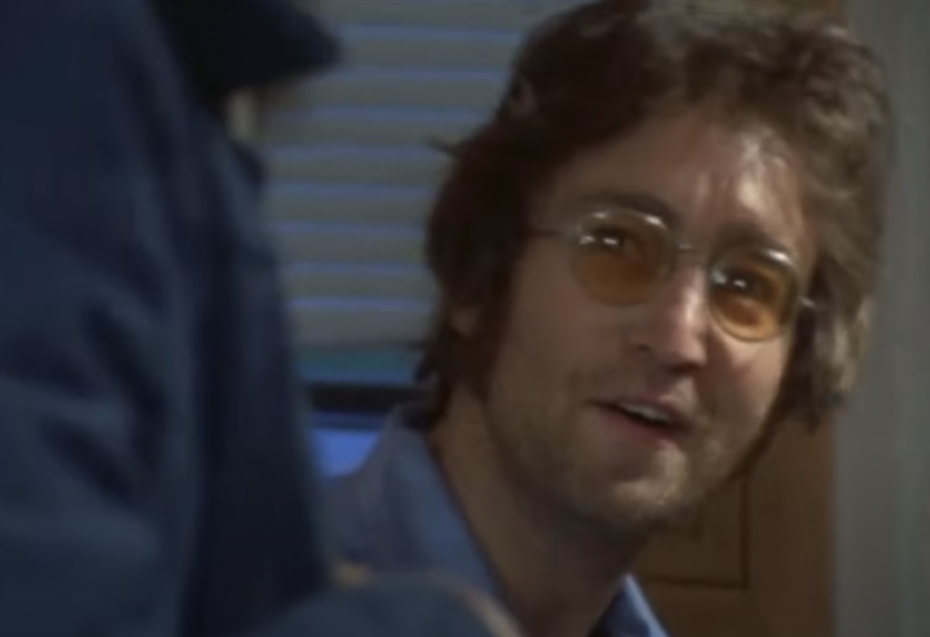 A close-up image of John Lennon in glasses being interviewed. 