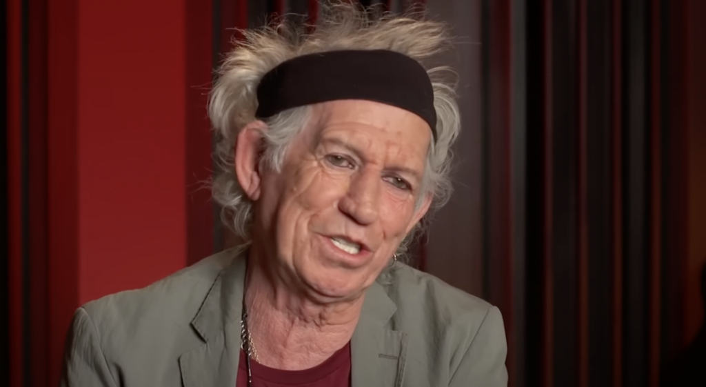 An older individual with gray, tousled hair and a black headband is speaking or being interviewed. They are wearing a light green jacket, a maroon shirt, and a necklace, set against a background with vertical dark stripes.