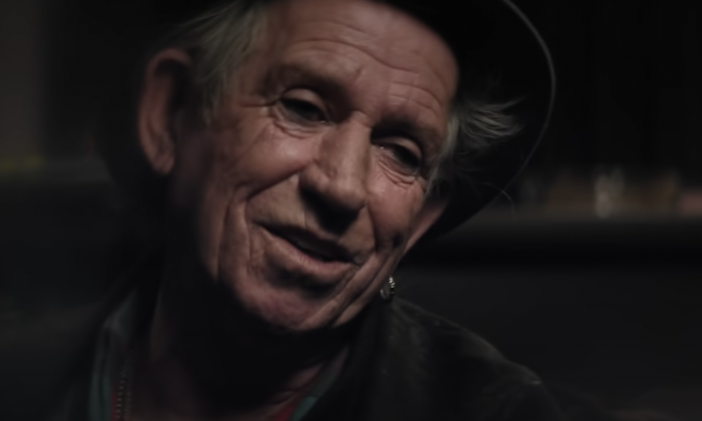 A person with a weathered face, grey hair, and a slight smile is wearing a black hat and a leather jacket. The background is dimly lit and out of focus.