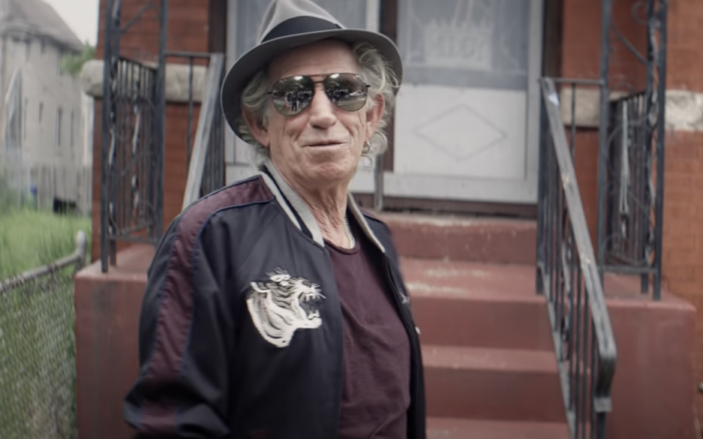 An older man with gray hair wearing a hat, sunglasses, navy jacket with dragon embroidery, and a dark shirt stands in front of a house with a staircase and metal railings. The background includes part of a red brick building and some greenery.