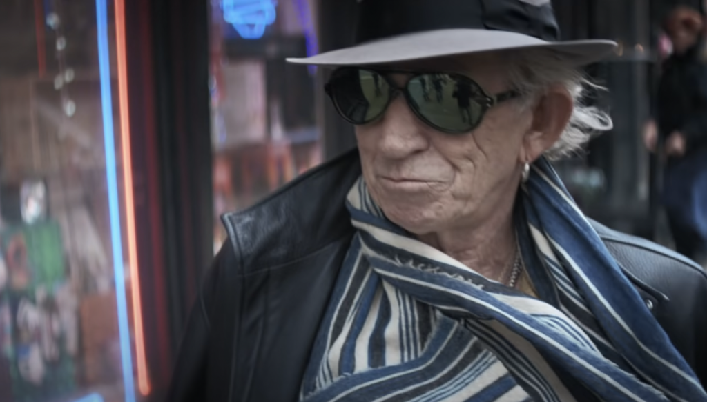 An older man wearing dark aviator sunglasses, a black hat, patterned blue and white scarf, and a leather jacket is standing in front of a storefront with neon lights. He has an earring in one ear and a slight smile.