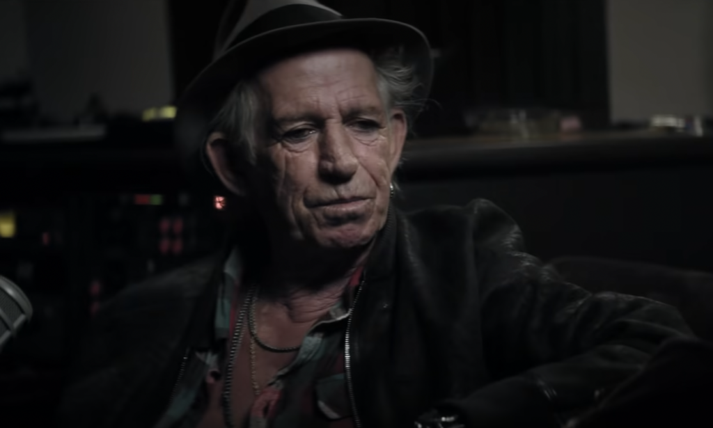 An older person with a weathered face, wearing a fedora hat and a leather jacket, sits in a dimly lit room, possibly a recording studio. The individual has a contemplative expression with visible recording equipment in the background.