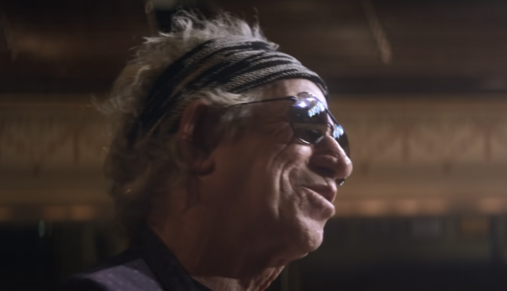 A person with disheveled gray hair and wearing a black and white patterned headband and sunglasses smiles. The background is out of focus, with warm lighting and an ornate design visible.