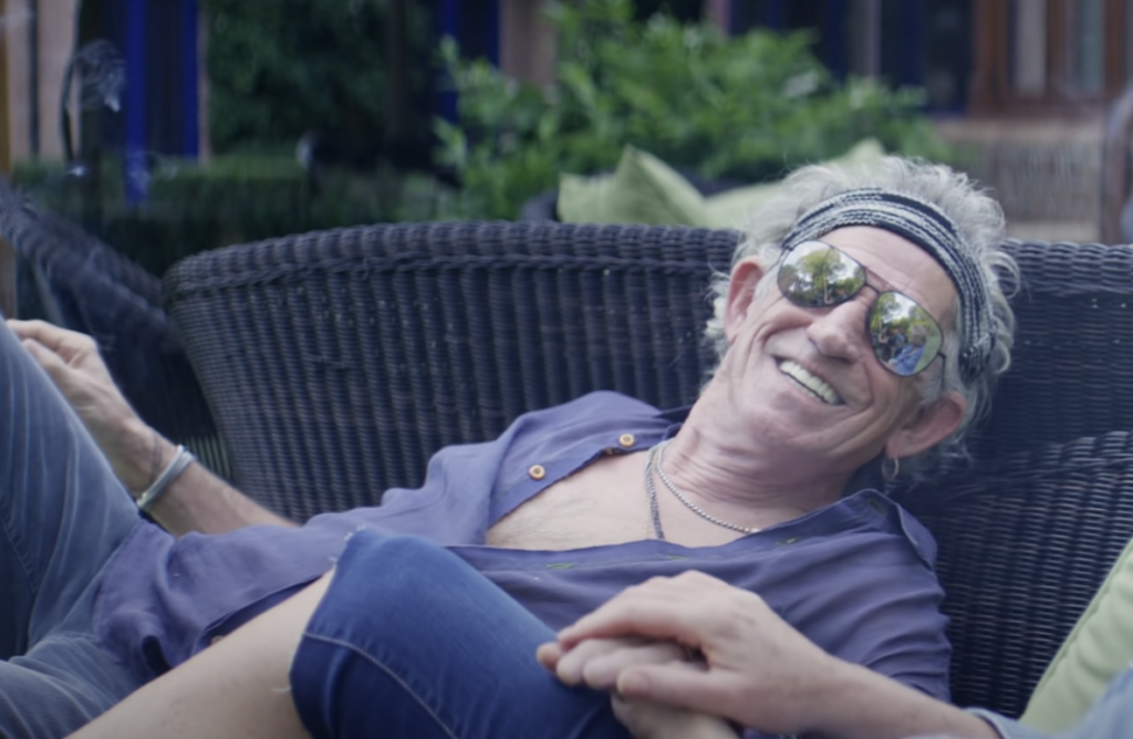 A man with gray hair, wearing sunglasses and a headband, relaxes on an outdoor wicker chair. He is smiling broadly and dressed casually in an open shirt and jeans. Lush greenery is in the background, suggesting a garden or park setting.