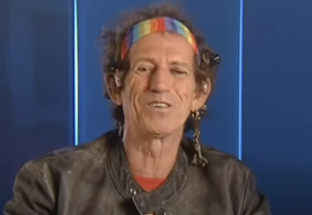 An older person with long, gray-brown hair tied back and wearing a rainbow-striped headband smiles at the camera. They are dressed in a black leather jacket, with blue background panels visible behind them.