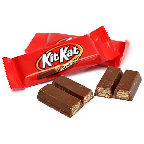Two KitKat bars in red wrappers with one wrapper opened, exposing the chocolate bars. Each chocolate bar is split into two pieces, revealing the layered, crispy wafer inside. The KitKat logo is visible on the wrappers.