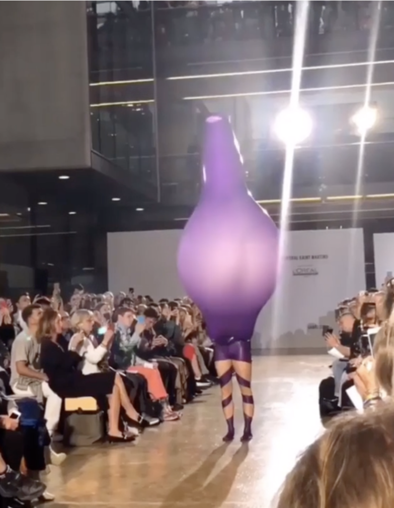 A fashion runway model walks in an outfit shaped like an oversized purple balloon, covering the entire body and head, with only legs visible. The audience, seated on both sides, observes and photographs the unique design. Bright lights illuminate the scene.