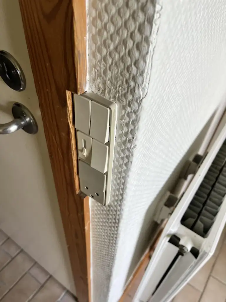 A light switch panel installed on a textured wall next to a wooden door frame. The switches are white, slightly askew, and one is missing its cover. Below the switches is an outlet. Part of a radiator is visible to the right.