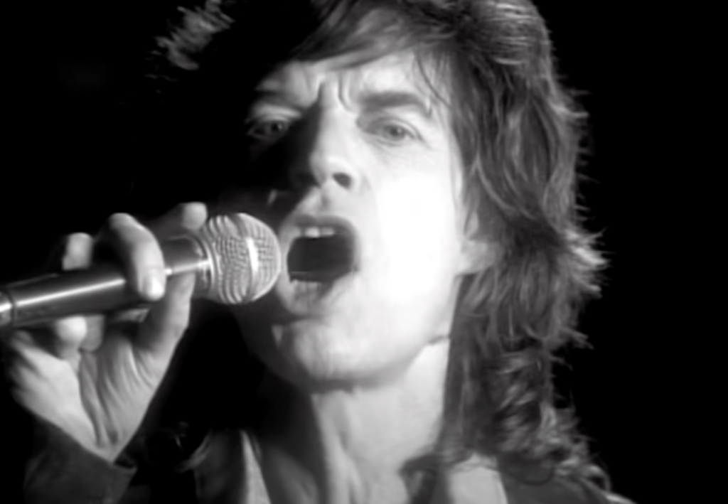 Black and white close-up image of a man with shoulder-length hair singing passionately into a microphone. His mouth is open wide, and his expression is intense. He is wearing a jacket over a shirt. The background is dark and out of focus.
