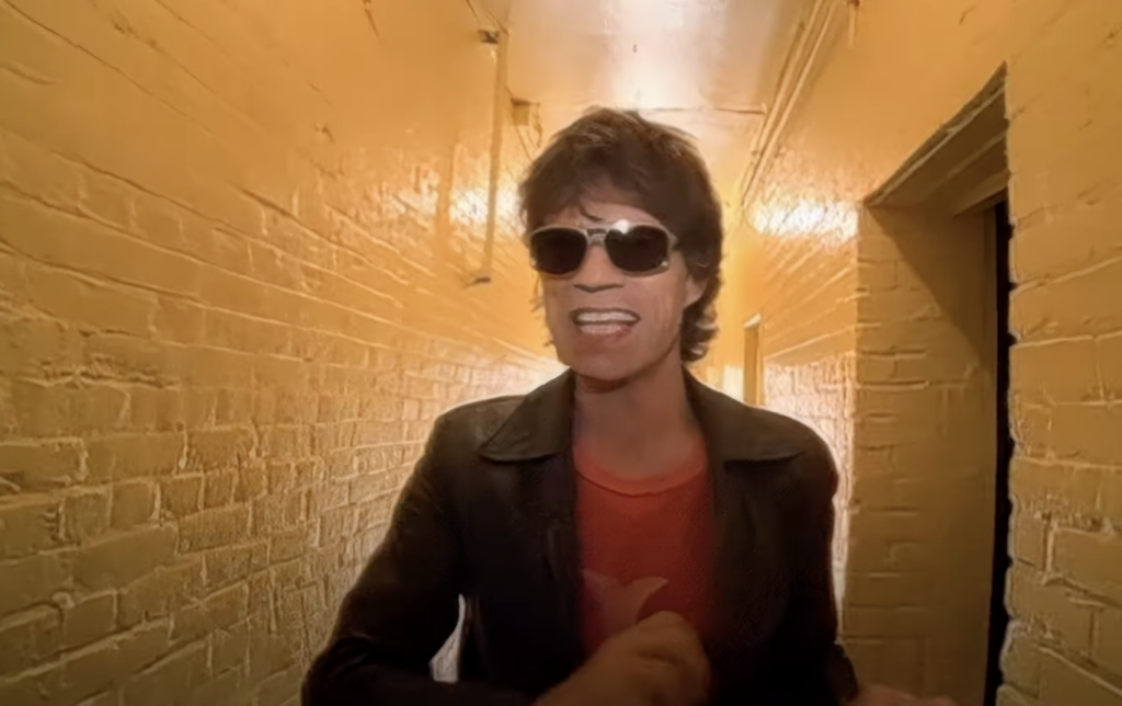 A person wearing sunglasses, a red shirt, and a dark jacket is standing in a narrow, dimly lit hallway with yellow-painted brick walls. The individual appears to be singing or talking animatedly.