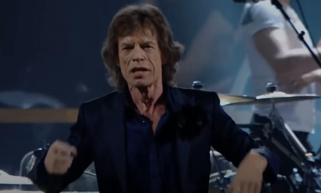 A man with long brown hair is performing on stage, wearing a dark jacket over a dark shirt. He appears to be singing or speaking into a microphone. In the background, a drummer is seen playing the drums. The stage is lit with blue lighting.
