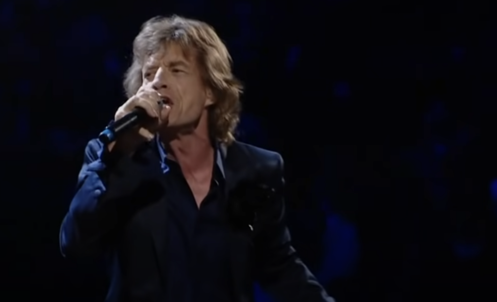 A person with long, wavy hair is passionately singing into a microphone on a dark stage. They are wearing a dark blazer over a shirt, and their expression is intense, conveying strong emotion. The background is dark, focusing attention on the performer.