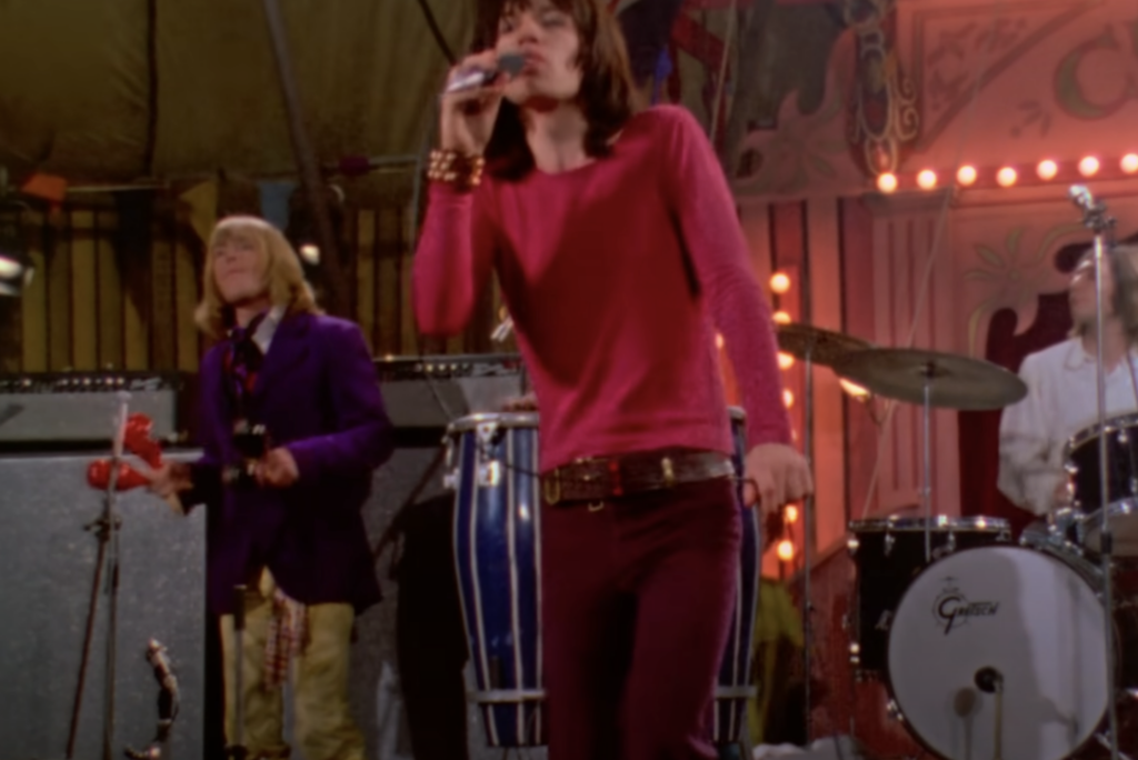 A singer in a red top and maroon pants performs passionately on stage, holding a microphone. A musician in a purple jacket plays a red maraca and percussion instruments in the background. A drummer in a white outfit plays on a drum set marked "Gretsch.