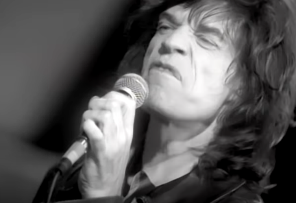 A black and white image of a person with long hair passionately singing into a microphone, eyes closed, wearing a leather jacket. The intense facial expression conveys emotion and energy.