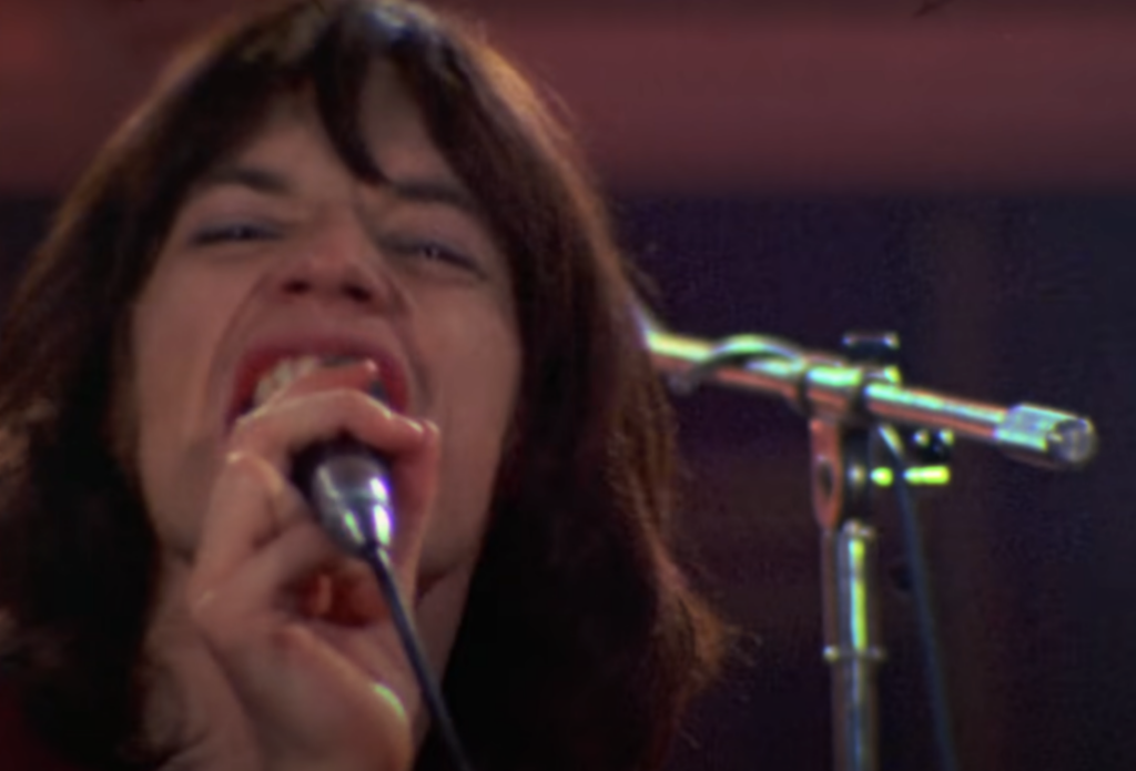 A person with shoulder-length dark hair is passionately singing into a microphone. Their mouth is open wide, and they are gripping the microphone with one hand. The background shows a metal microphone stand and blurred stage lights.