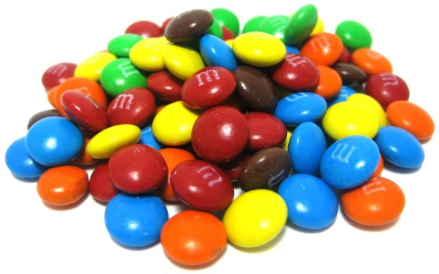 A pile of colorful candy-coated chocolate pieces, primarily red, yellow, blue, brown, green, and orange. Some candies have white "m" imprints on them. The candies are round and glossy, creating a vibrant and playful assortment.