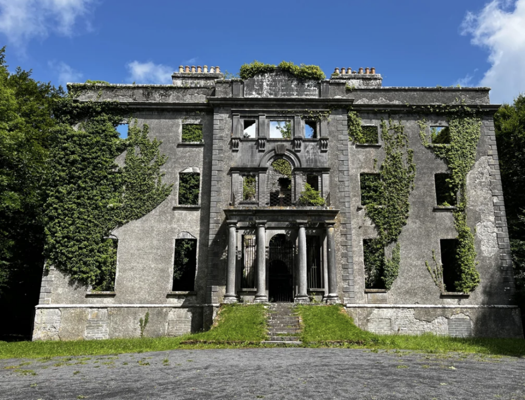 A large, abandoned building stands with overgrown ivy climbing its walls. The worn facade, broken windows, and missing roof sections reflect its dilapidated state. The entrance features a stone portico with pillars. Blue sky and greenery add contrast.