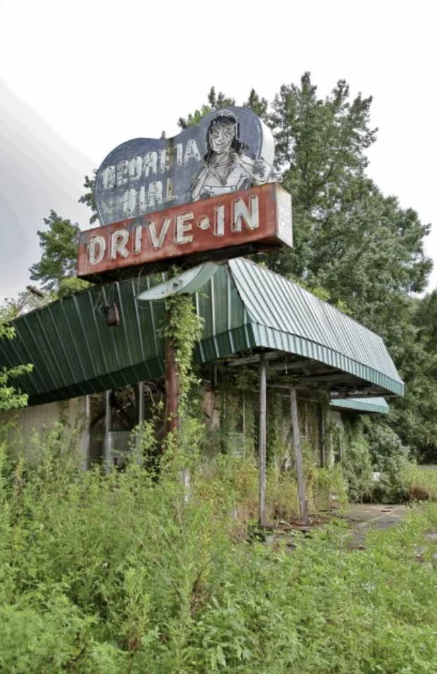 An overgrown, abandoned drive-in restaurant with a worn-out sign reading "GEORGIA GIRL DRIVE IN" on top. The sign features a faded image of a woman, and the building is surrounded by dense greenery and foliage.