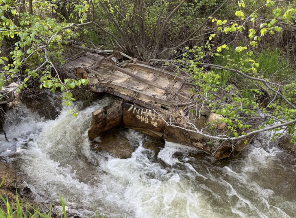 A rusty, overturned car chassis lies in a small, flowing creek surrounded by greenery. The vehicle is partially submerged in the water, with its deteriorated metal frame and axles visible. Lush trees and bushes line the creek's edge.