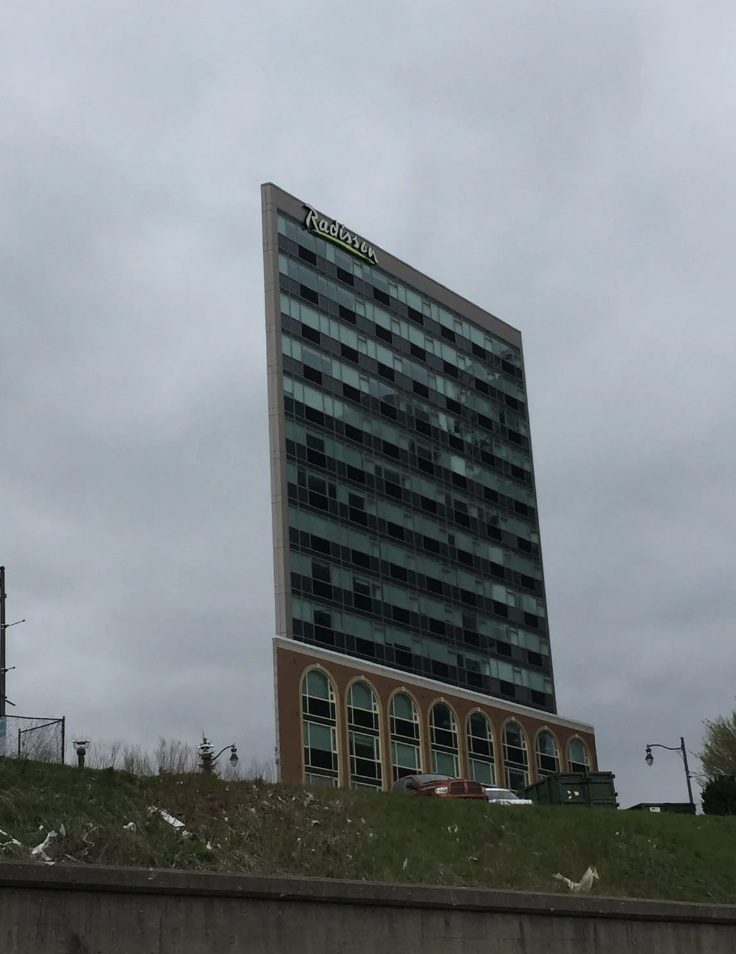 A tall building with a sign "Radisson" at the top stands against a cloudy sky. The structure features a mix of glass windows and brick at the base, with multiple arches on the lower floors. Sparse greenery and a concrete barrier are seen in the foreground.