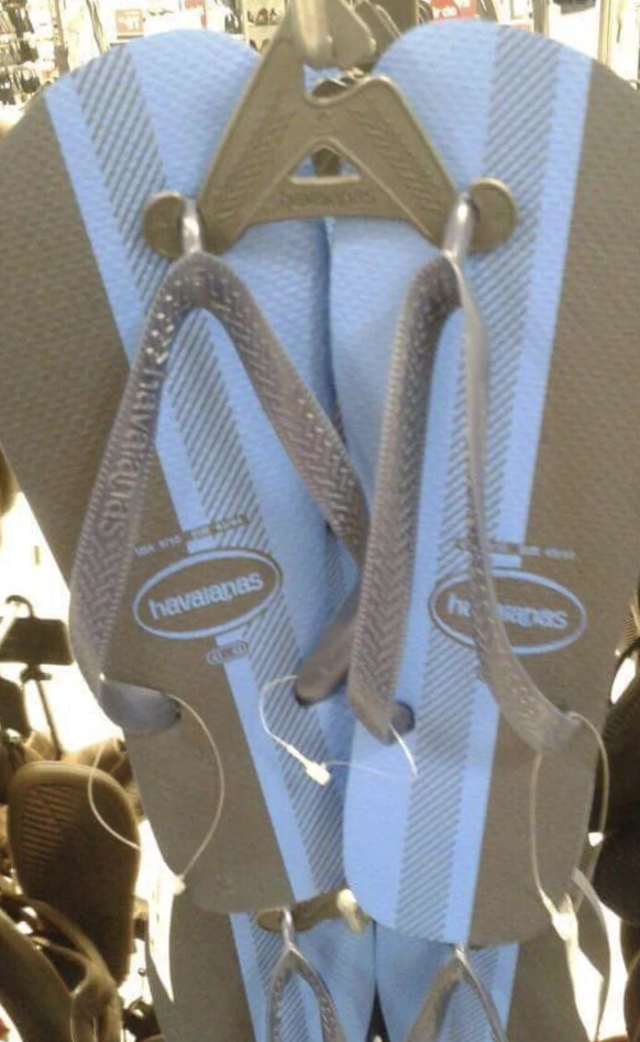 A pair of Havaianas flip-flops with a blue and gray striped design hanging on a display rack in a store. The straps and sole feature the brand’s logo. Other flip-flops in various colors and styles can be seen in the background.